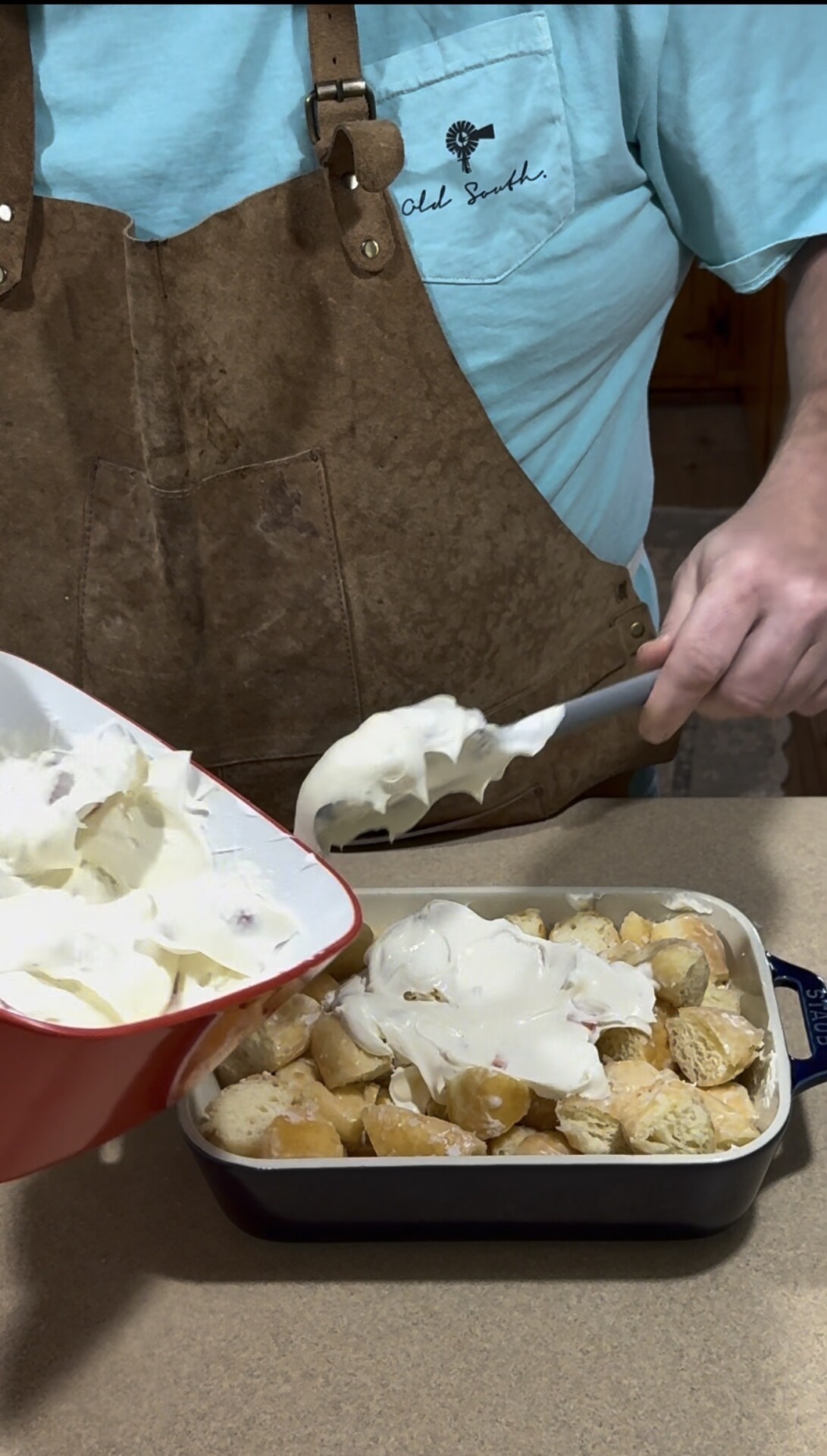 A person in a blue shirt and brown apron spreads whipped topping over a dish filled with pieces of bread or cake using a spatula.