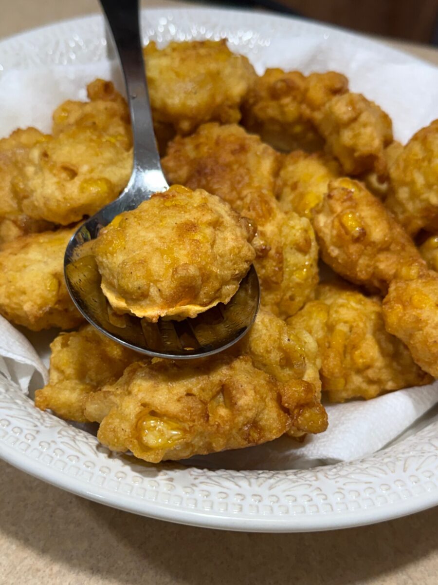 A close-up of golden-brown corn fritters in a white bowl lined with paper towels, with a spoon lifting one fritter.