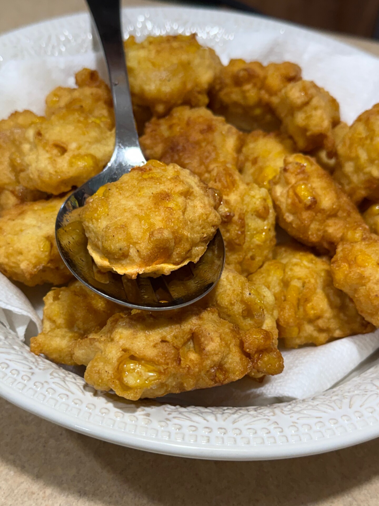 A close-up of golden-brown corn fritters in a white bowl lined with paper towels, with a spoon lifting one fritter.
