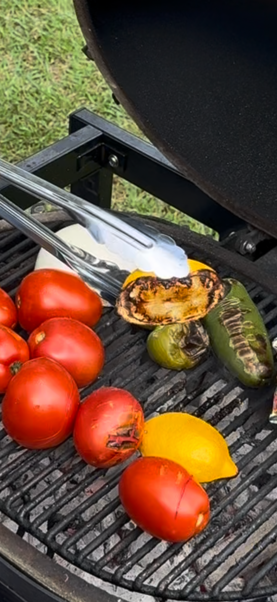 A grill with fresh tomatoes, a charred yellow squash, a lemon wedge, a green pepper, and metal tongs over the open flame. The grill is set outdoors on green grass.