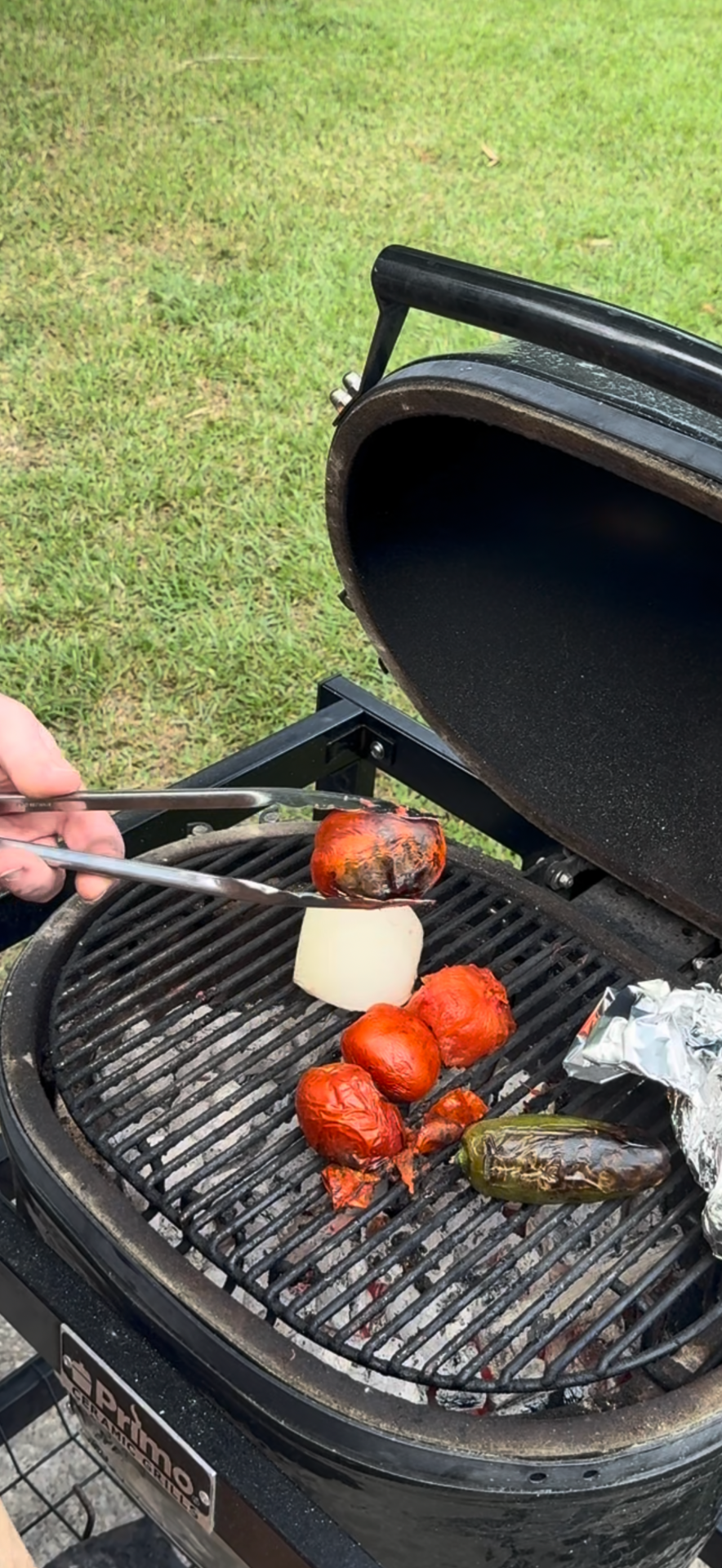 A hand uses tongs to grill tomatoes, onion, and peppers on a round charcoal grill outside, with grass visible in the background.