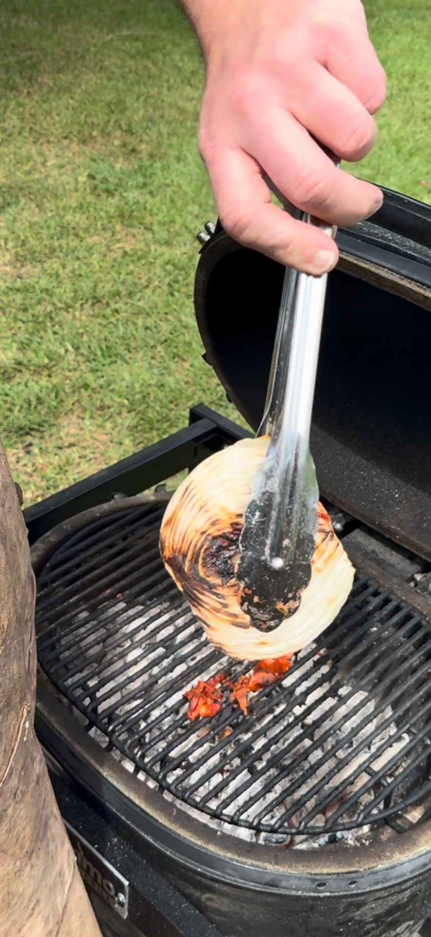 A hand uses metal tongs to hold a charred onion half over a grill with a black lid outdoors on green grass.