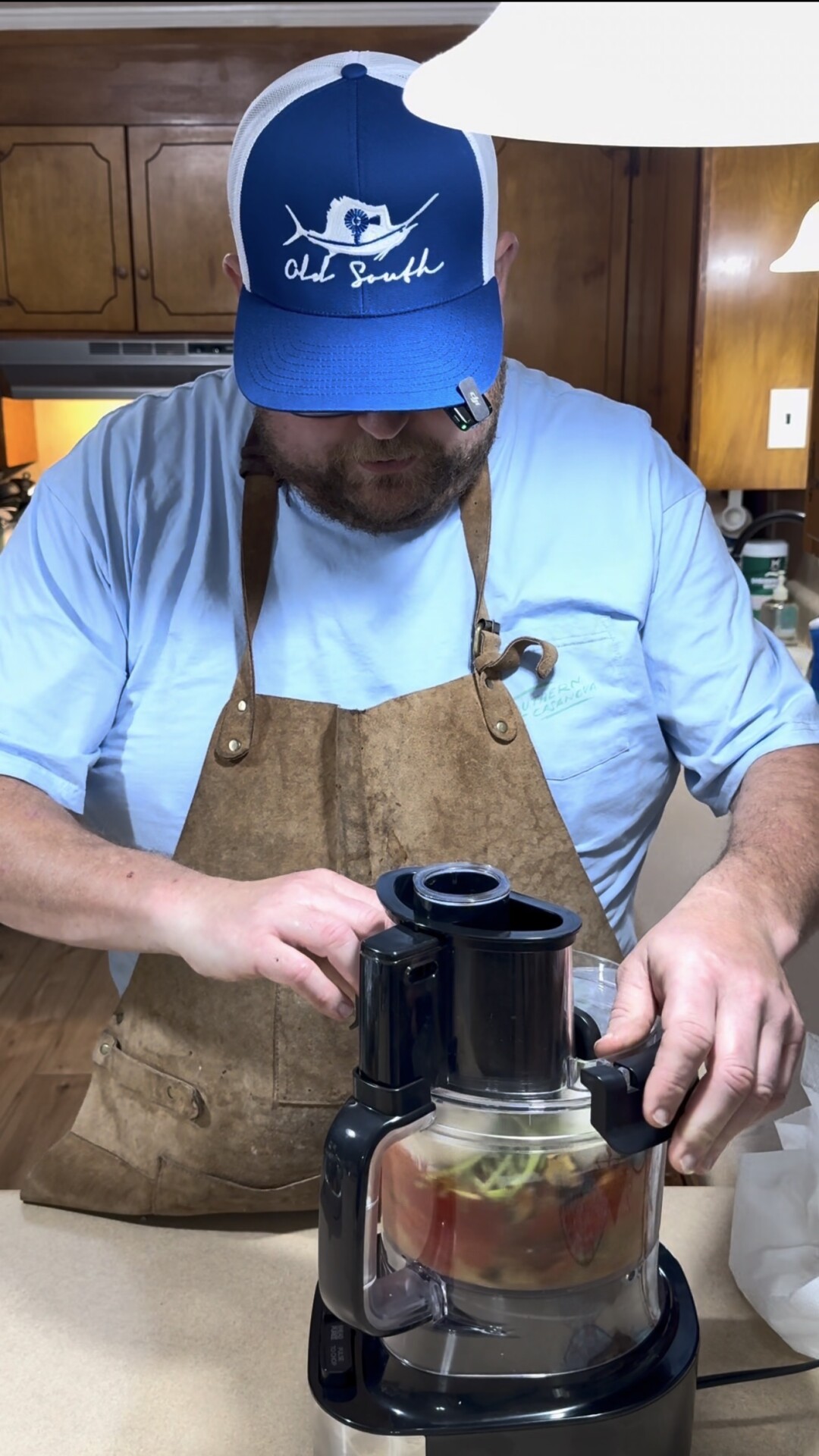 A man in a blue cap, blue shirt, and brown apron uses a juicer in a kitchen, pressing fruit or vegetables into the machine.