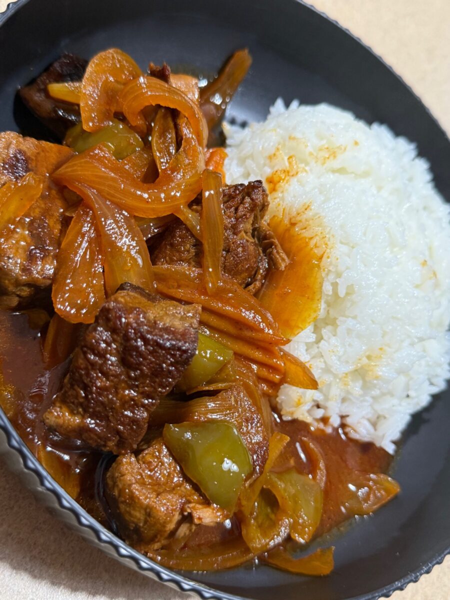 A bowl with white rice and a savory stew containing braised meat, sliced onions, green bell peppers, and a rich brown sauce.