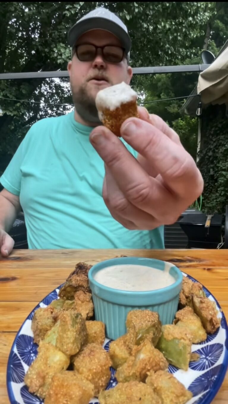 A person in a teal shirt holds up a piece of fried food dipped in sauce. A plate with more fried pieces and a ramekin of dipping sauce sits on a wooden table outdoors. Trees and umbrellas are in the background.
