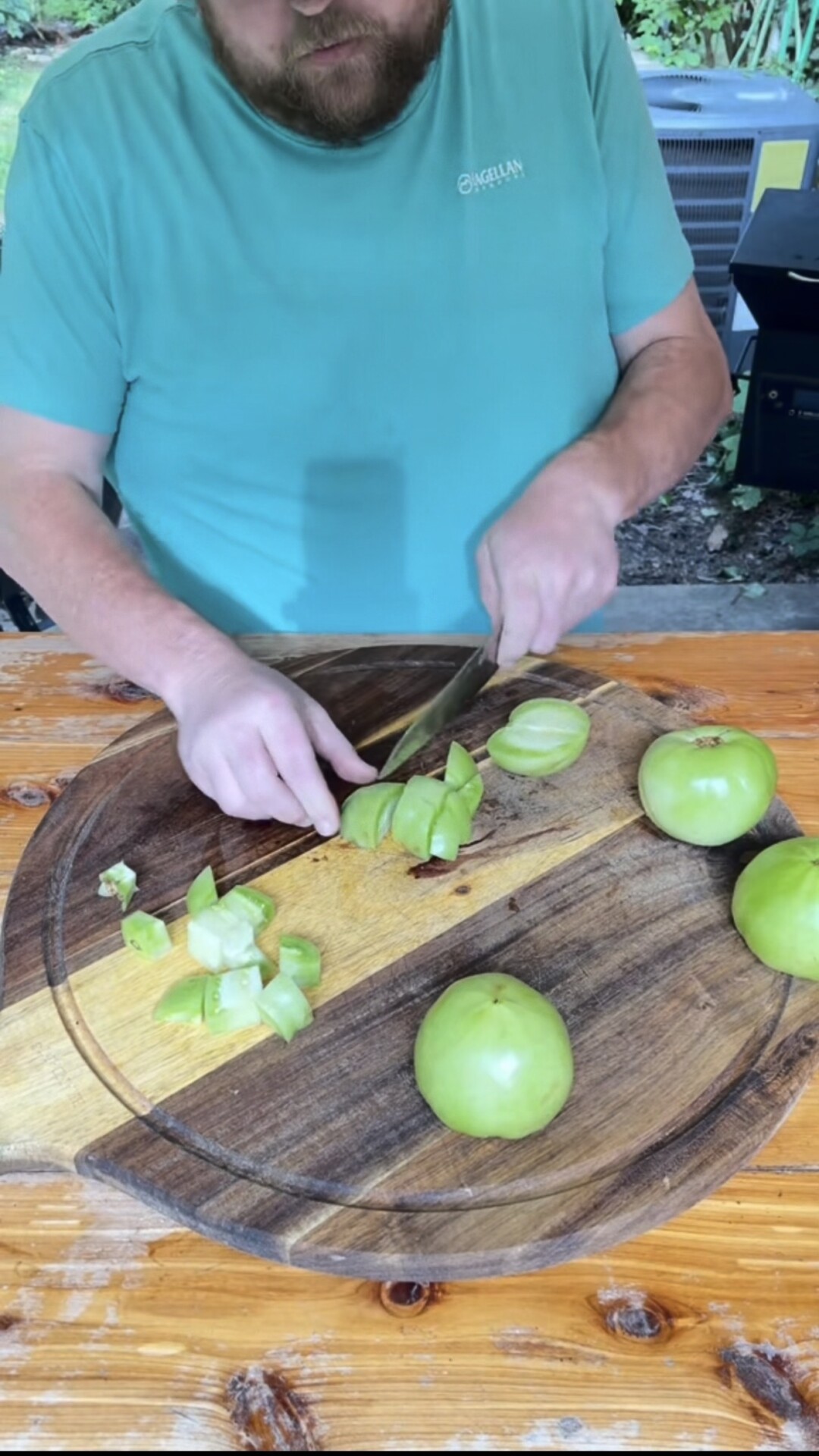 A person in a teal shirt slices green tomatoes on a wooden cutting board outdoors, with whole and chopped tomatoes visible.