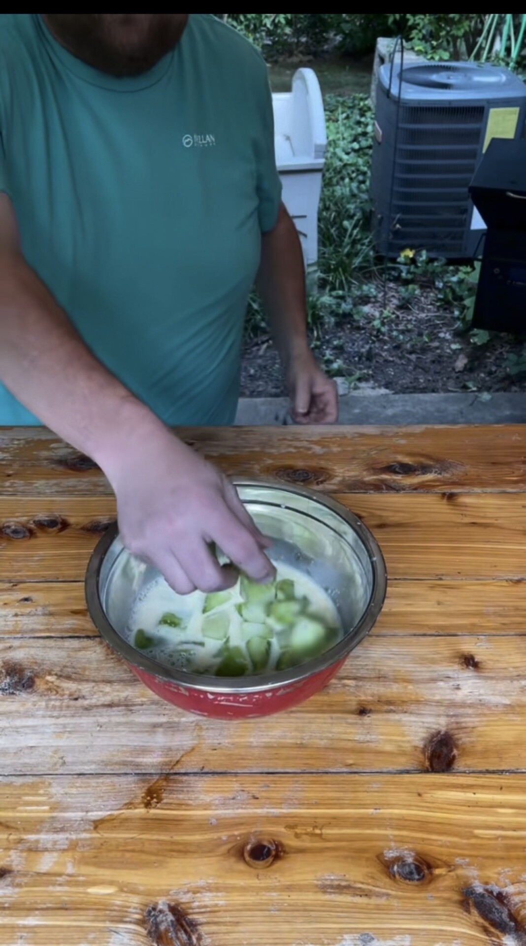 A person in a teal shirt is dipping cucumber slices into a metal bowl filled with liquid on a wooden table outdoors.