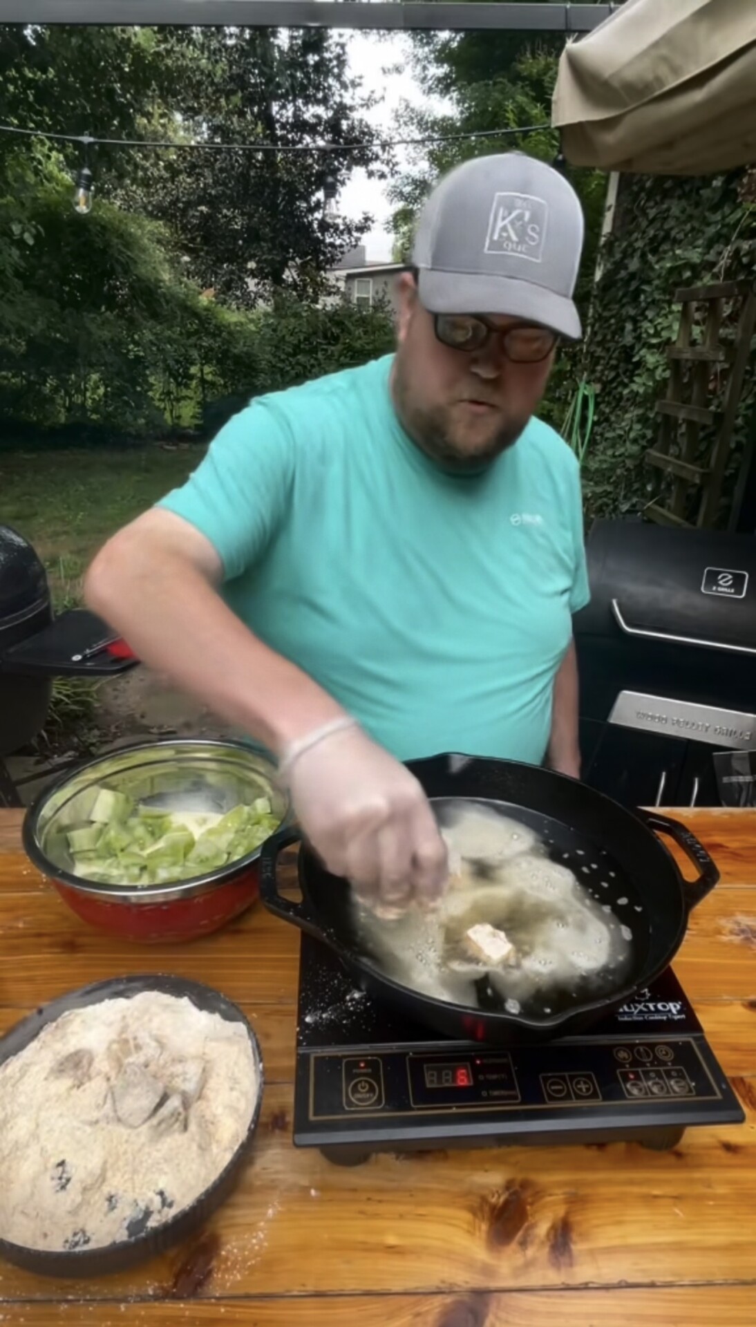 A man in a teal shirt and gray cap fries battered food outdoors on an electric stove, with bowls of vegetables and batter nearby, surrounded by greenery.