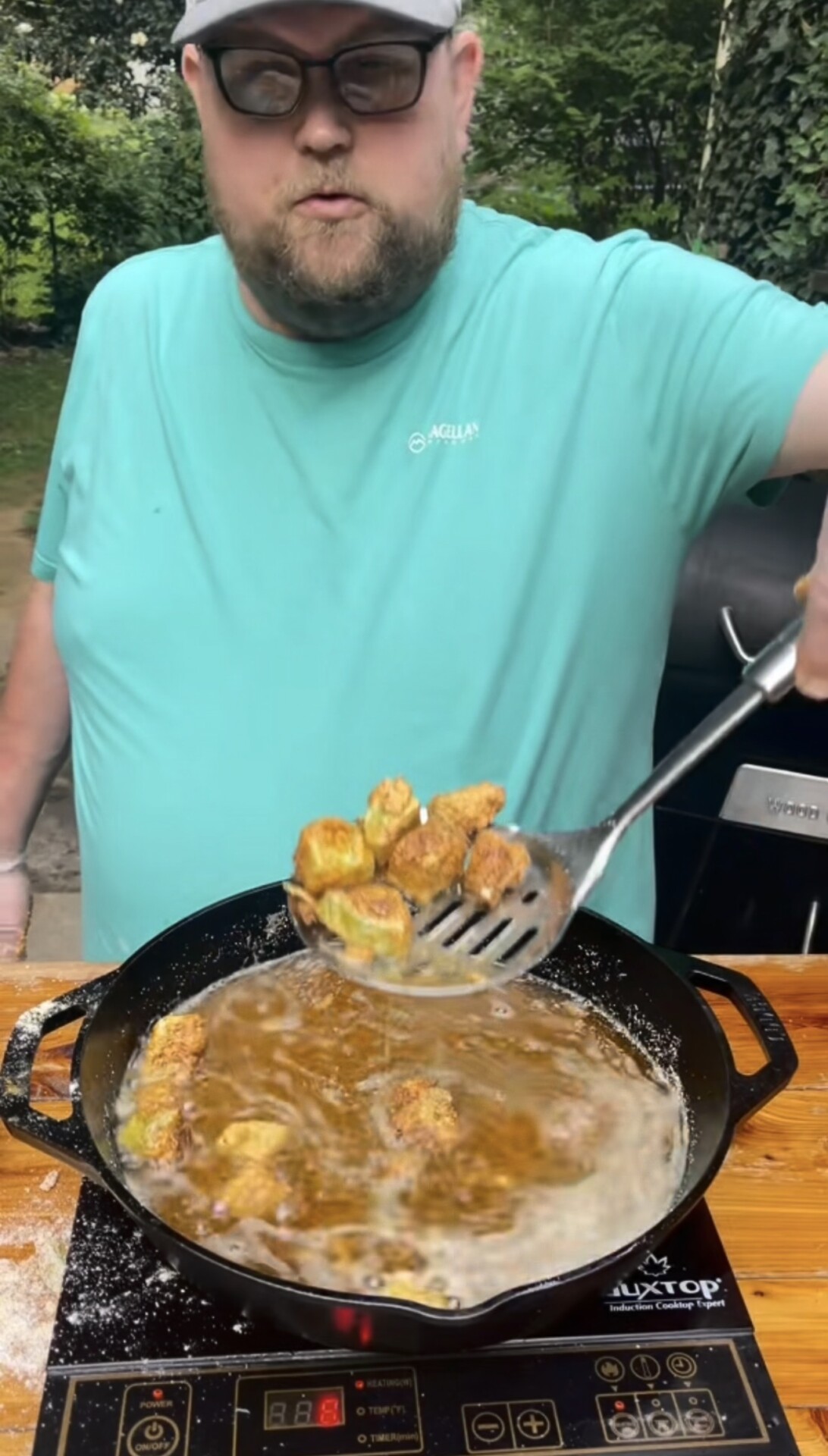 A person in a teal shirt fries battered food in a cast iron skillet outdoors, using a slotted spatula to lift pieces from bubbling oil.