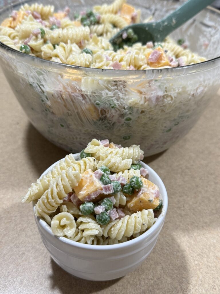 A small white bowl filled with rotini pasta salad containing peas, cubed cheese, and diced ham, with a larger bowl of the same salad in the background.