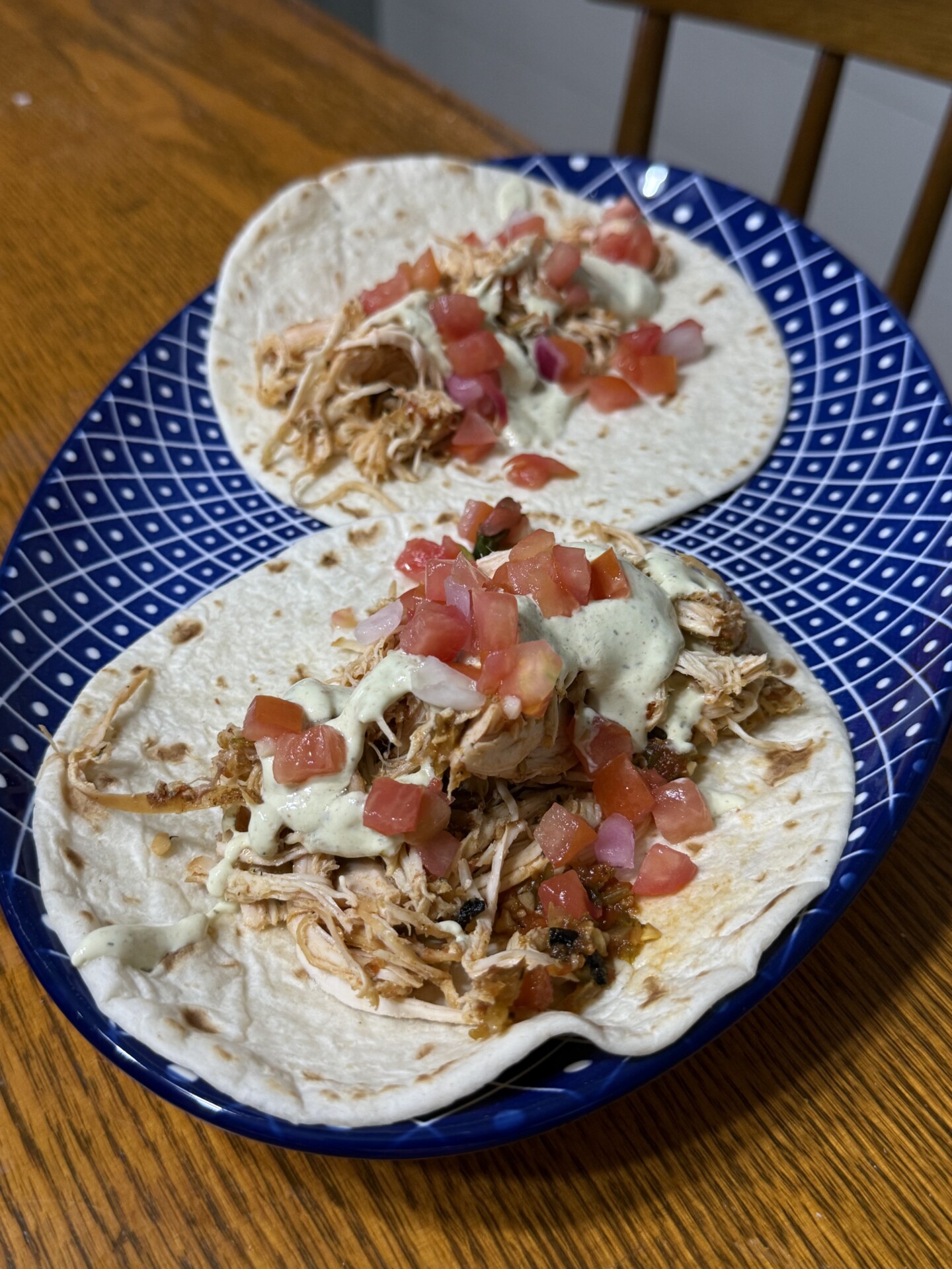 Two shredded chicken tacos on flour tortillas, topped with diced tomatoes and creamy sauce, served on a blue patterned plate on a wooden table.