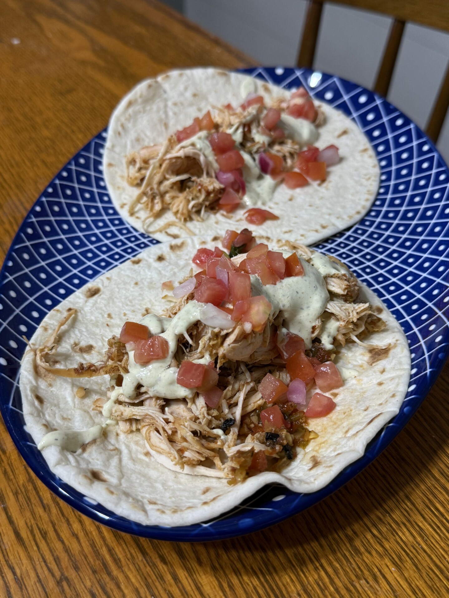 Two flour tortillas filled with shredded chicken, diced tomatoes, and a drizzle of creamy sauce, served on a blue patterned plate atop a wooden table.