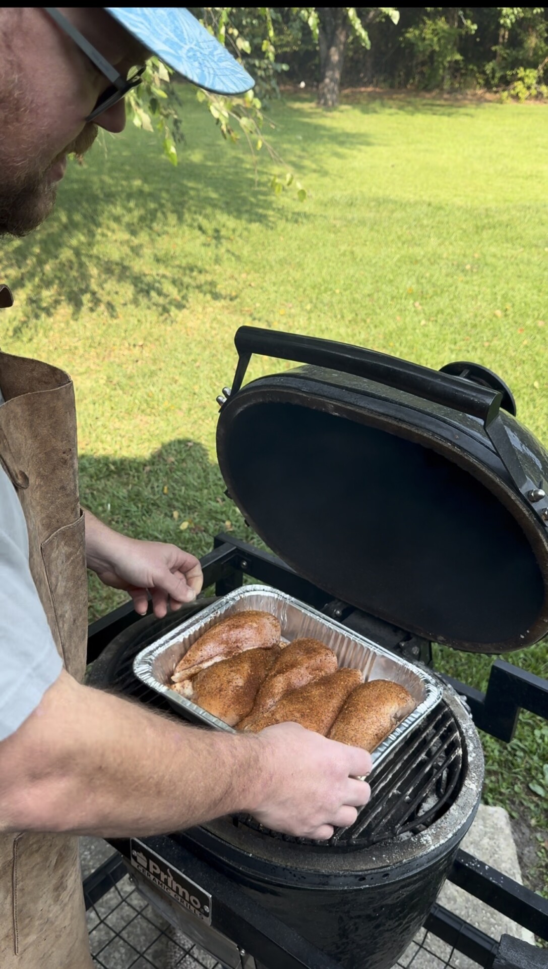 A person wearing an apron places a tray of seasoned chicken breasts on an outdoor grill with a domed lid. Lush green grass and trees are visible in the background.