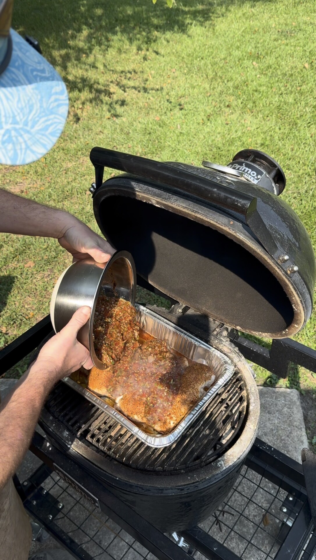 A person pours marinade from a metal bowl onto meat in a foil tray on a black charcoal grill outdoors, with green grass and a blue patterned object in the background.