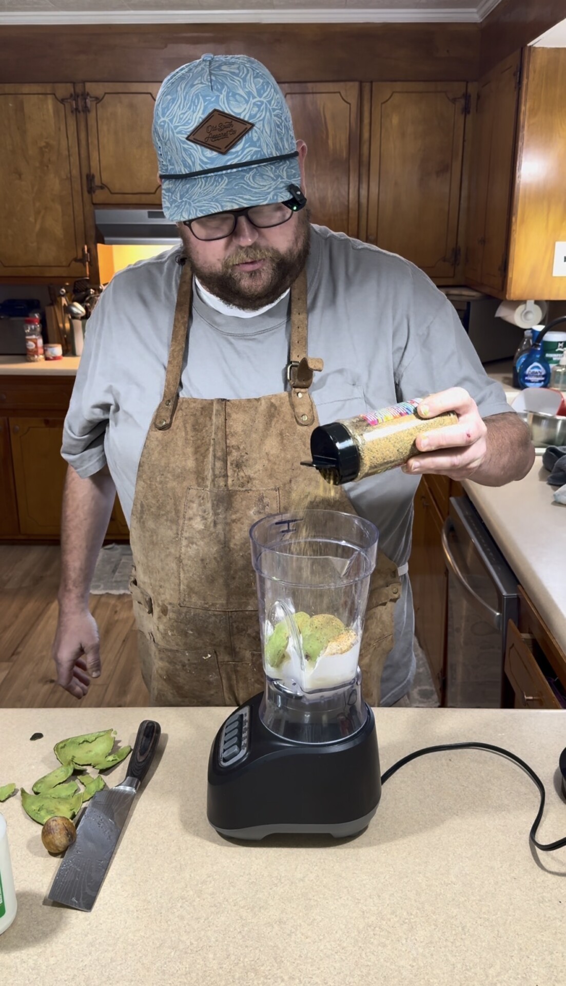 A person wearing an apron and blue cap pours seasoning into a blender filled with ingredients in a kitchen, with a knife and avocado on the counter.
