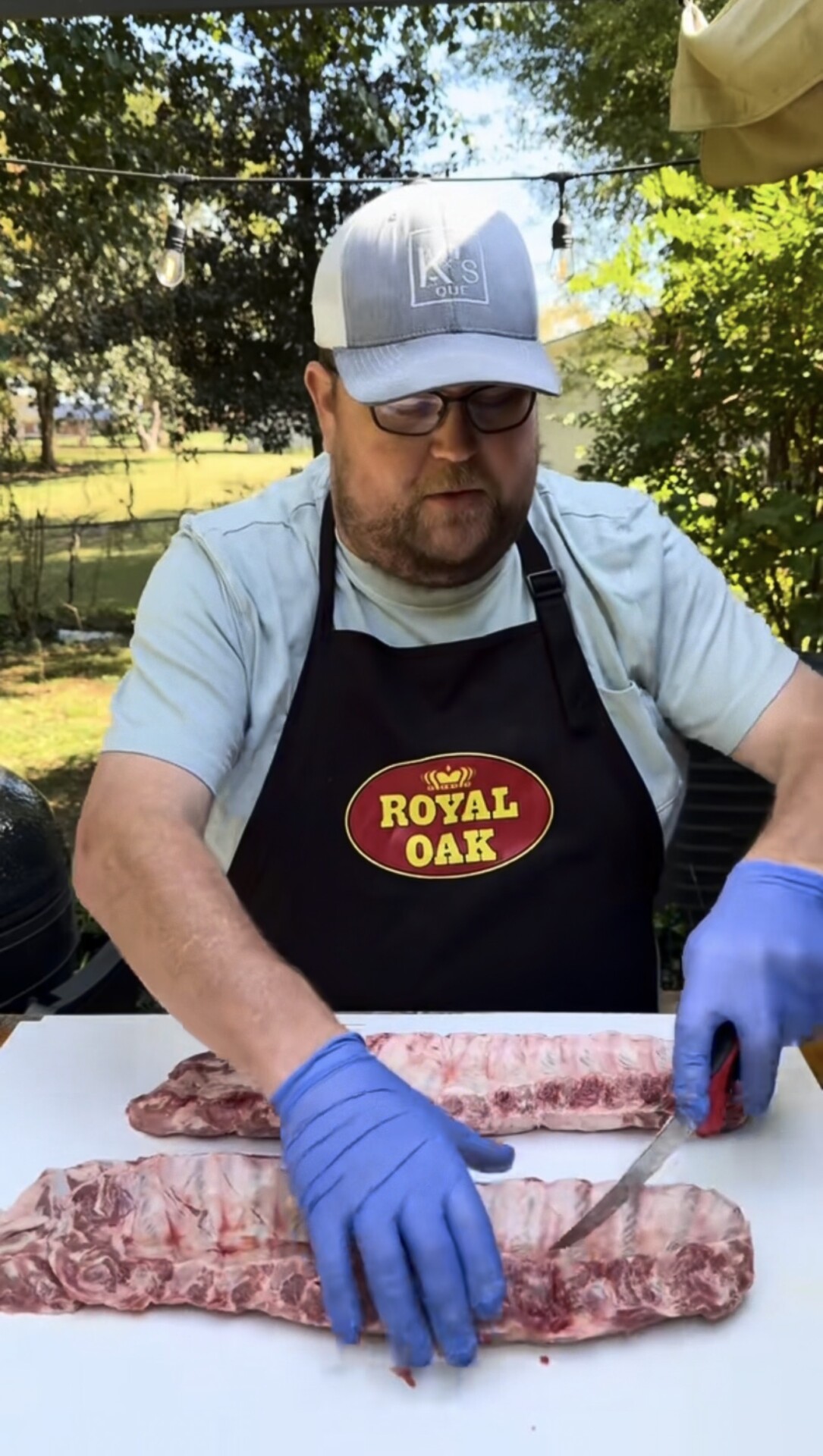 A man in a Royal Oak apron and blue gloves slices raw ribs on a white table outdoors, with trees and a lawn in the background.