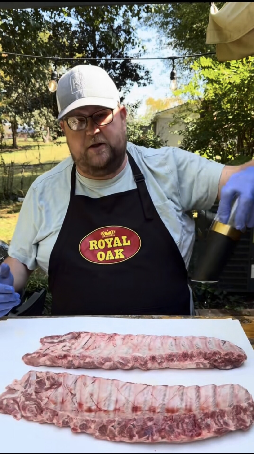 A man wearing a Royal Oak apron and blue gloves prepares to season two raw racks of ribs on a table outdoors. Trees, string lights, and a yard are visible in the background.