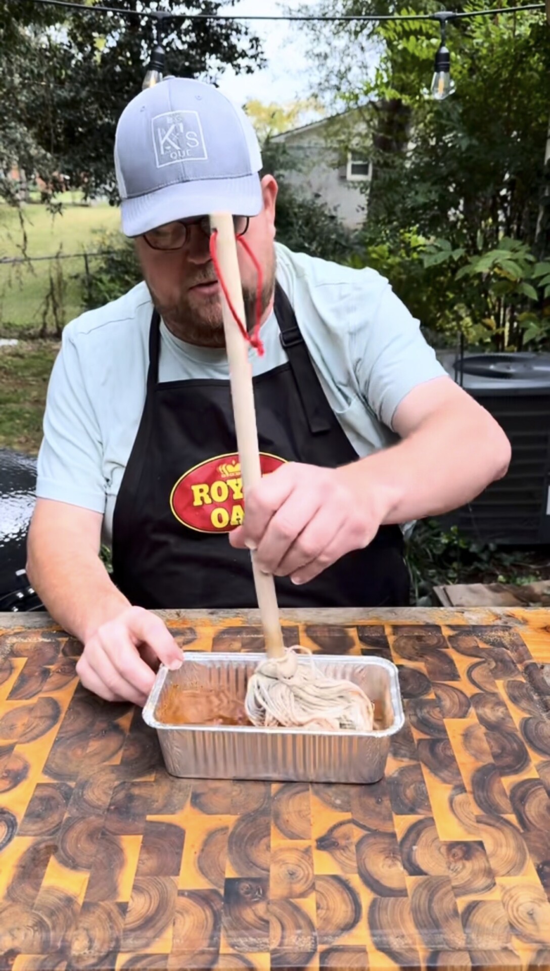 A person in an apron stirs a pan of brown food with a wooden stick attached to string, outdoors on a patterned wooden table with trees and a house in the background.