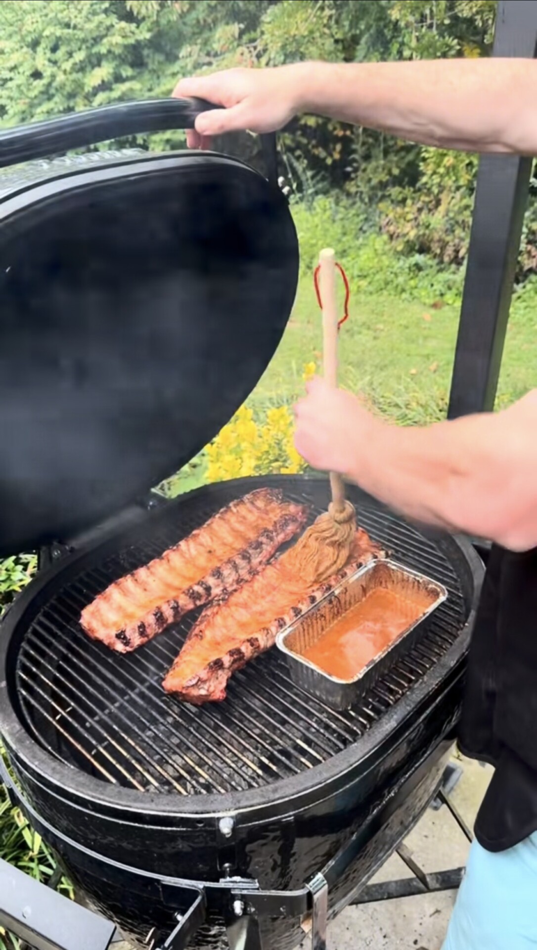 Person basting two racks of ribs with sauce on a grill using a mop brush. There is a tray of sauce next to the ribs, and smoke is rising from the grill. Lush greenery is visible in the background.