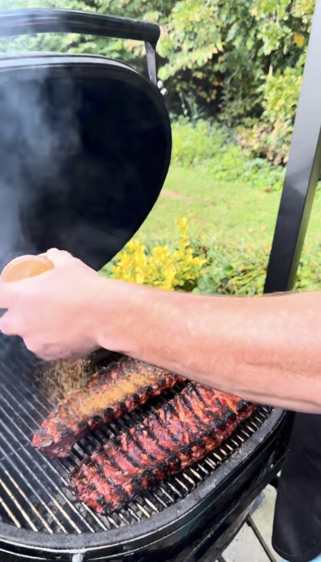 A person sprinkles seasoning onto ribs cooking on a grill, with smoke rising and a green garden in the background.