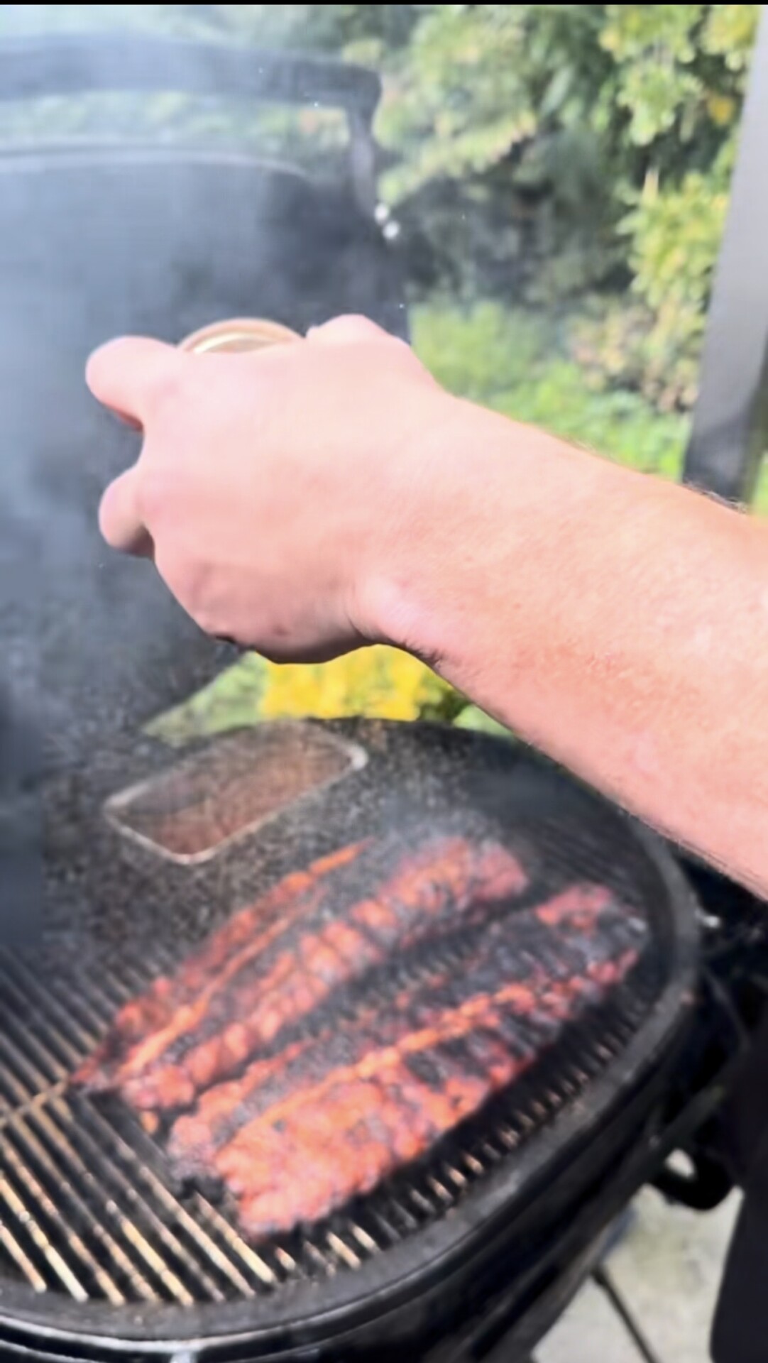 A hand sprinkles seasoning over ribs cooking on a smoky grill outdoors, with green foliage visible in the background.
