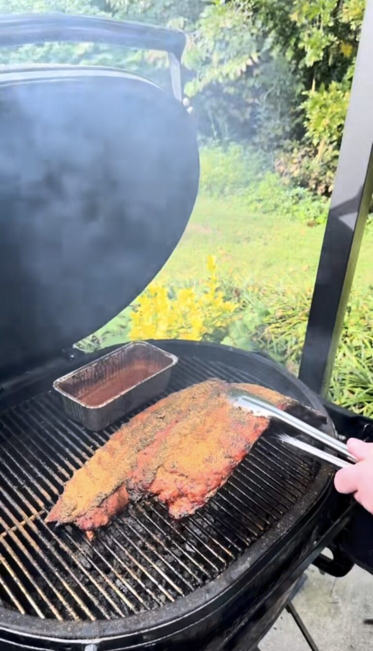 A person uses tongs to grill seasoned ribs on a barbecue smoker outdoors, with a small tray of sauce nearby and green foliage in the background.