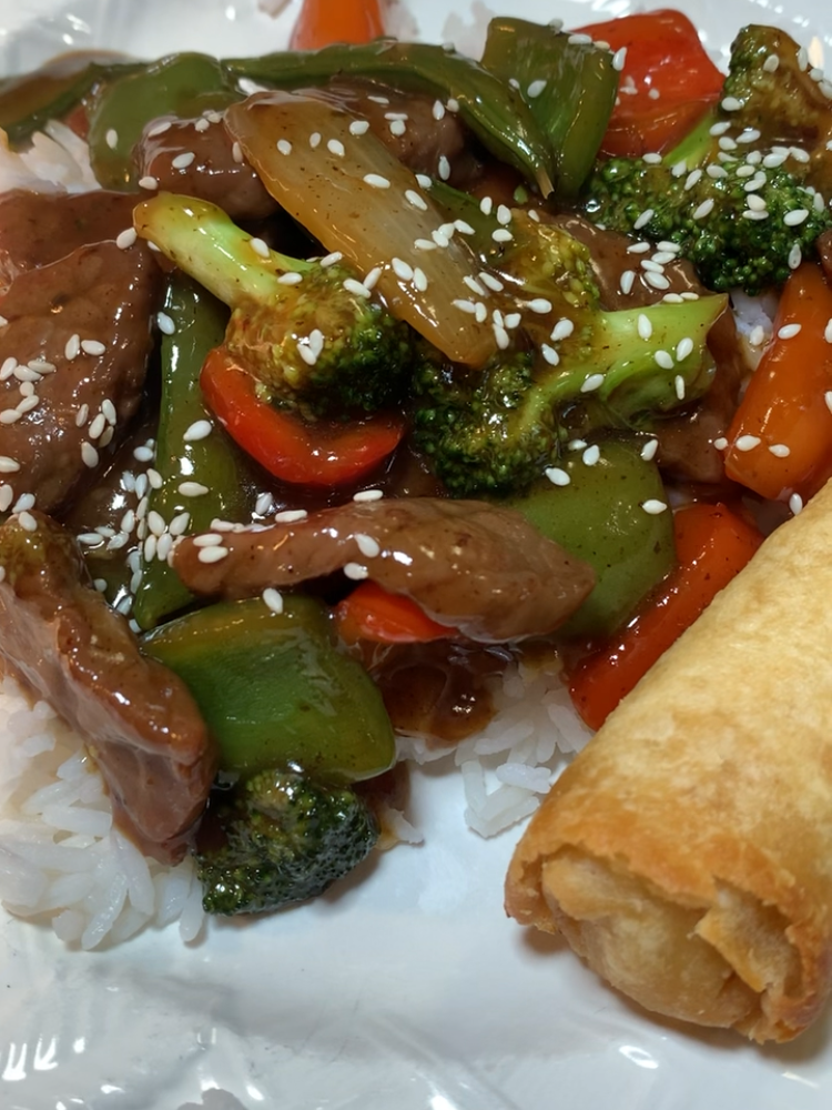 Beef stir-fry with broccoli, bell peppers, carrots, and onions in sauce over white rice, topped with sesame seeds, served with a golden egg roll on a white plate.