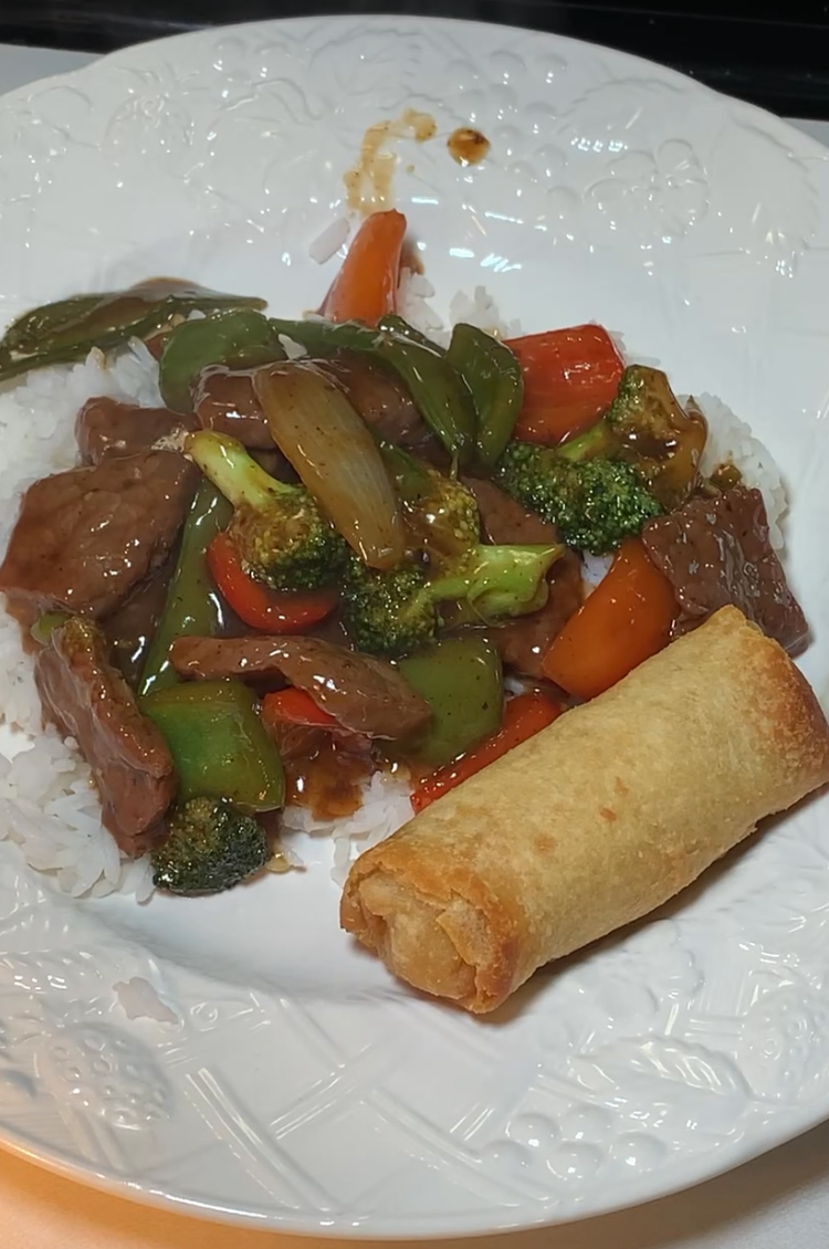 A plate of beef stir-fry with bell peppers, broccoli, onions, and carrots served over white rice, alongside a fried egg roll.