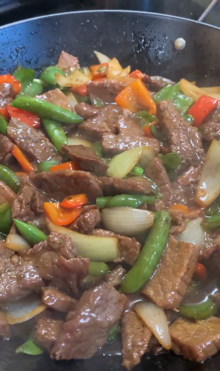 Beef stir-fry with colorful bell peppers, snap peas, celery, and onions cooking in a pan, coated in a glossy sauce.