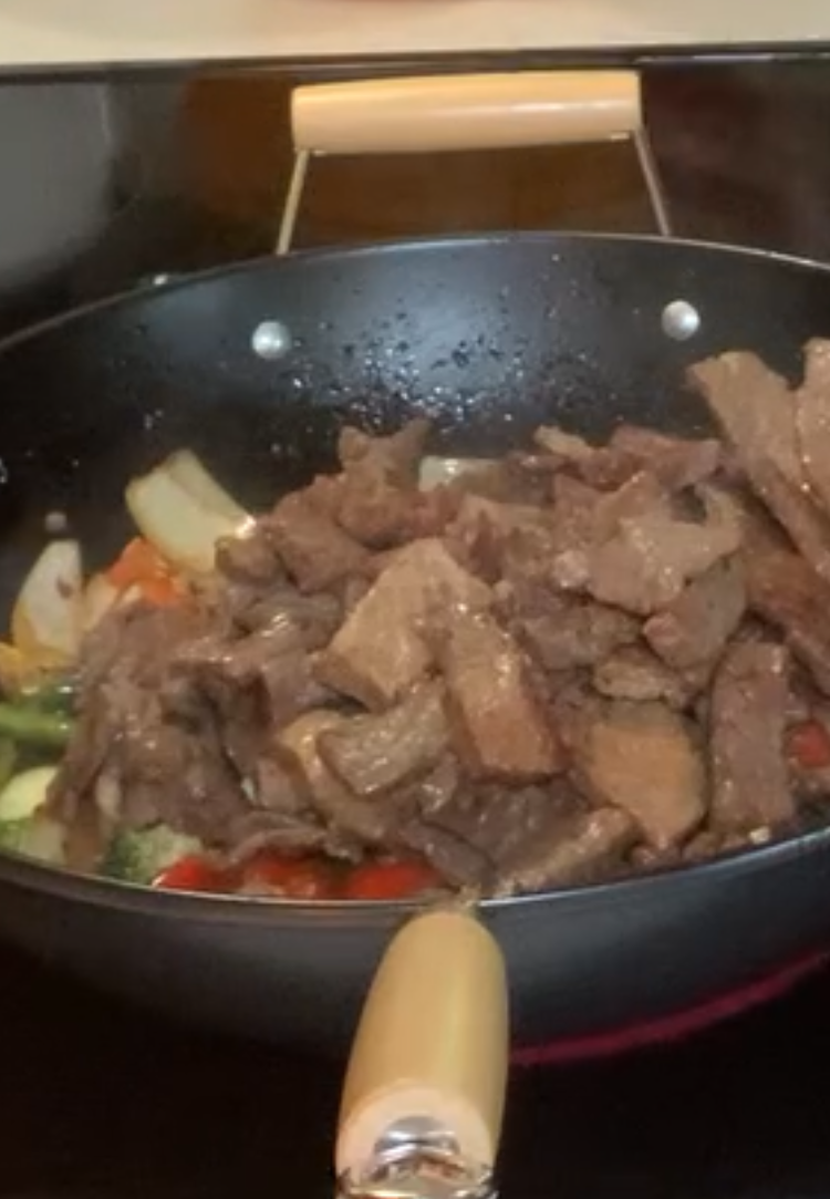 A black wok on a stove contains cooked sliced beef, onions, and assorted vegetables like broccoli and bell peppers, ready for stirring.