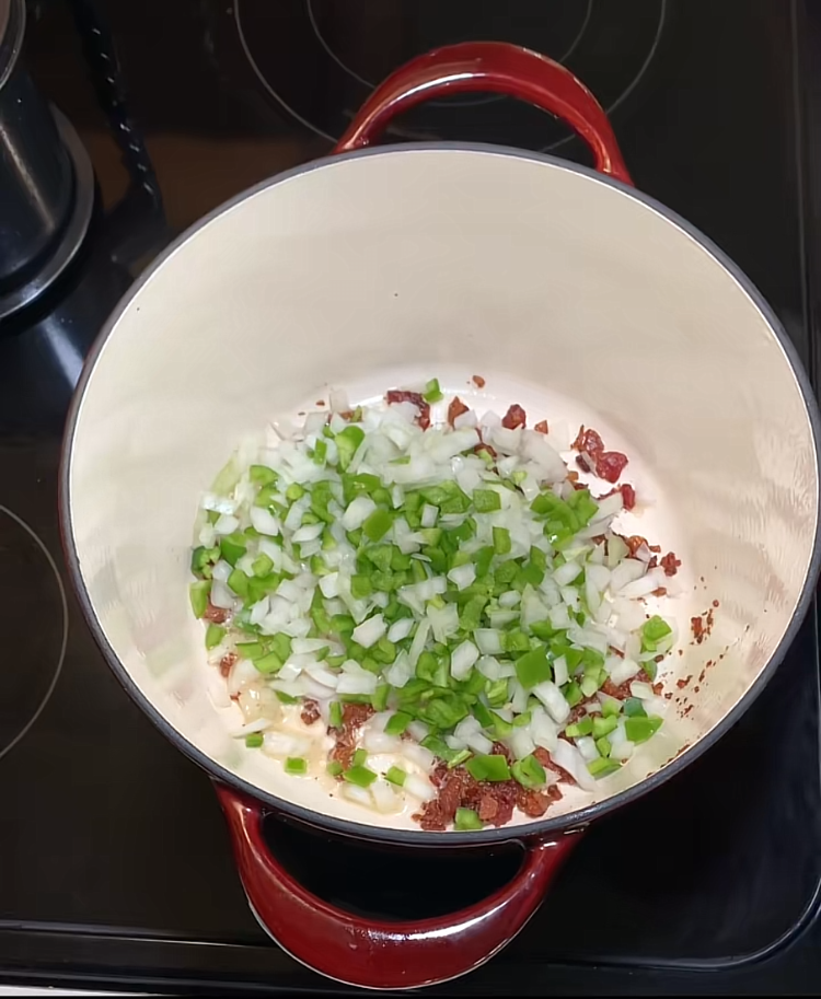 A red Dutch oven on a stovetop contains chopped green bell pepper, onions, and browned bits, likely from cooking bacon or meat, being sautéed together.