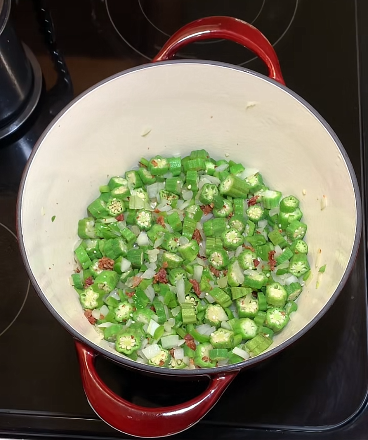 A red Dutch oven on a stovetop contains chopped okra, onions, and tomatoes being sautéed.