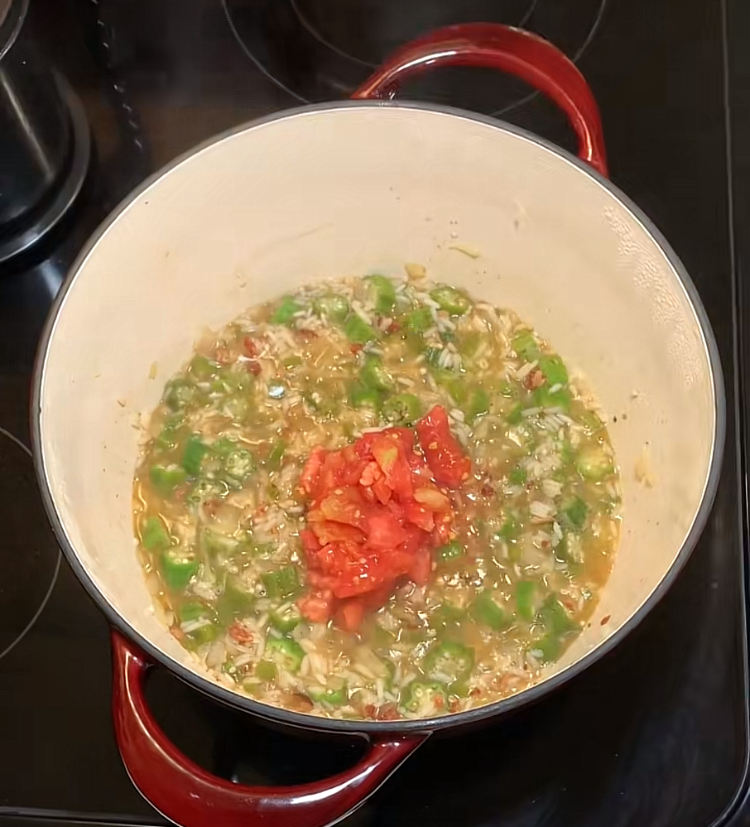 A red-handled pot on a stovetop contains chopped green peppers, onions, and fresh tomatoes cooking together.