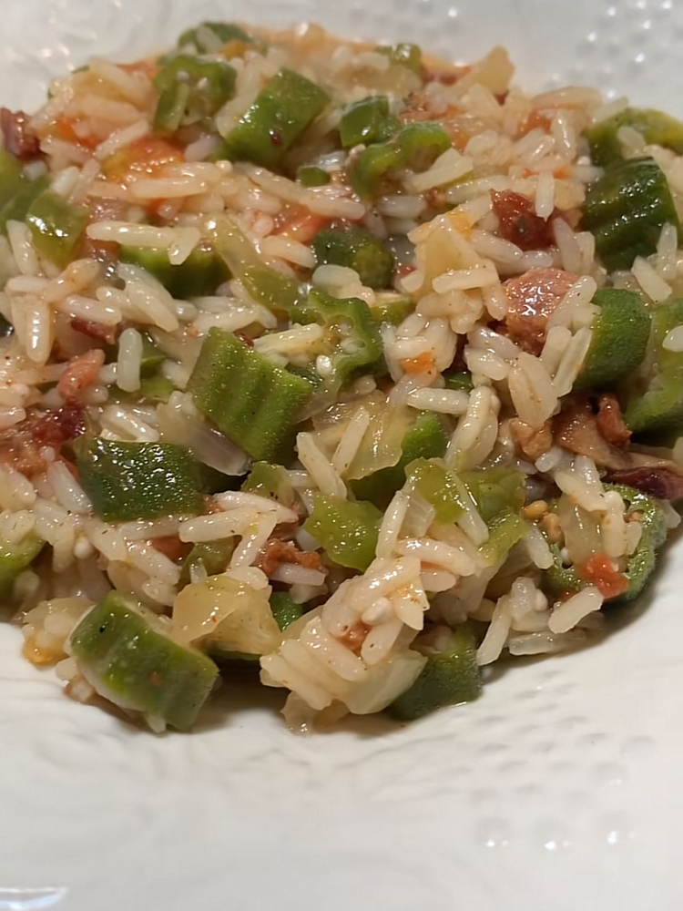 A close-up of a dish featuring white rice mixed with chopped okra, tomatoes, and small pieces of bacon, served in a white bowl.