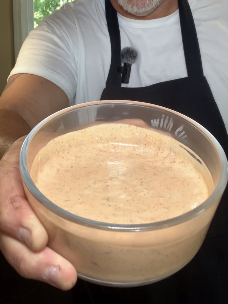A person wearing a black apron and white shirt holds up a glass bowl filled with a creamy, orange-colored sauce or dip.