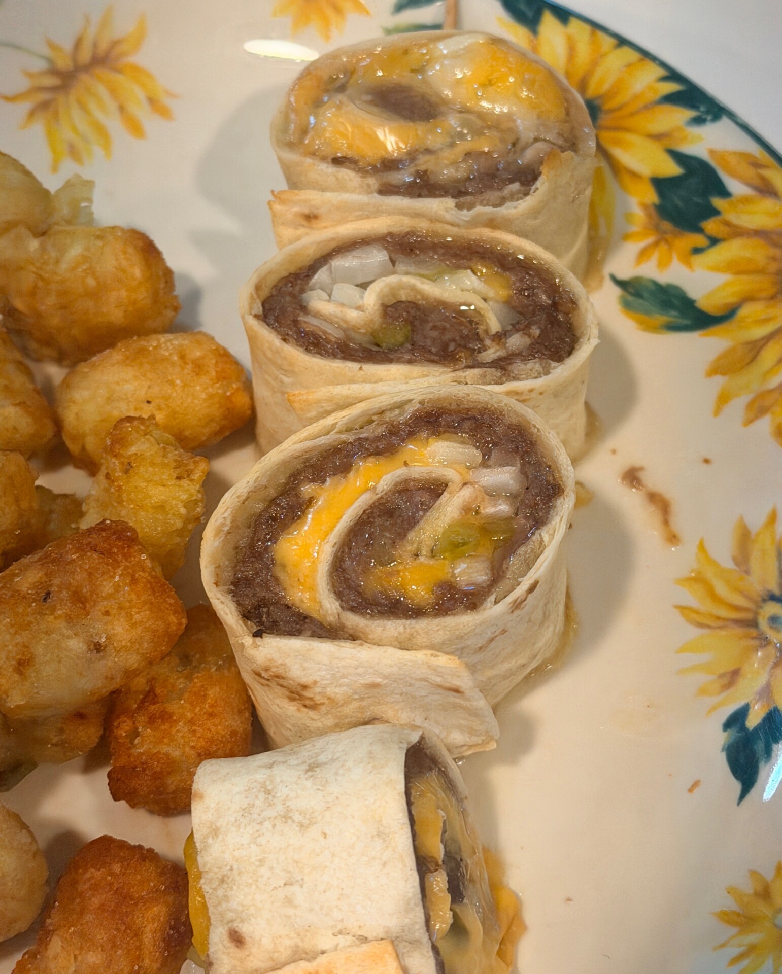 A close-up of four beef and cheese pinwheels made with tortillas, served alongside crispy tater tots on a sunflower-patterned plate.
