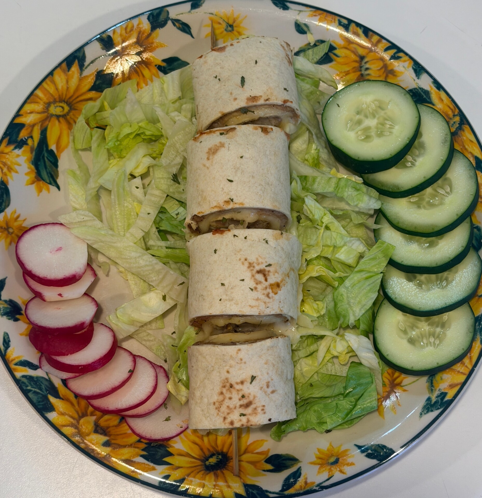 A floral plate with a sliced wrap on shredded lettuce, next to sliced cucumbers and radishes arranged on the side.