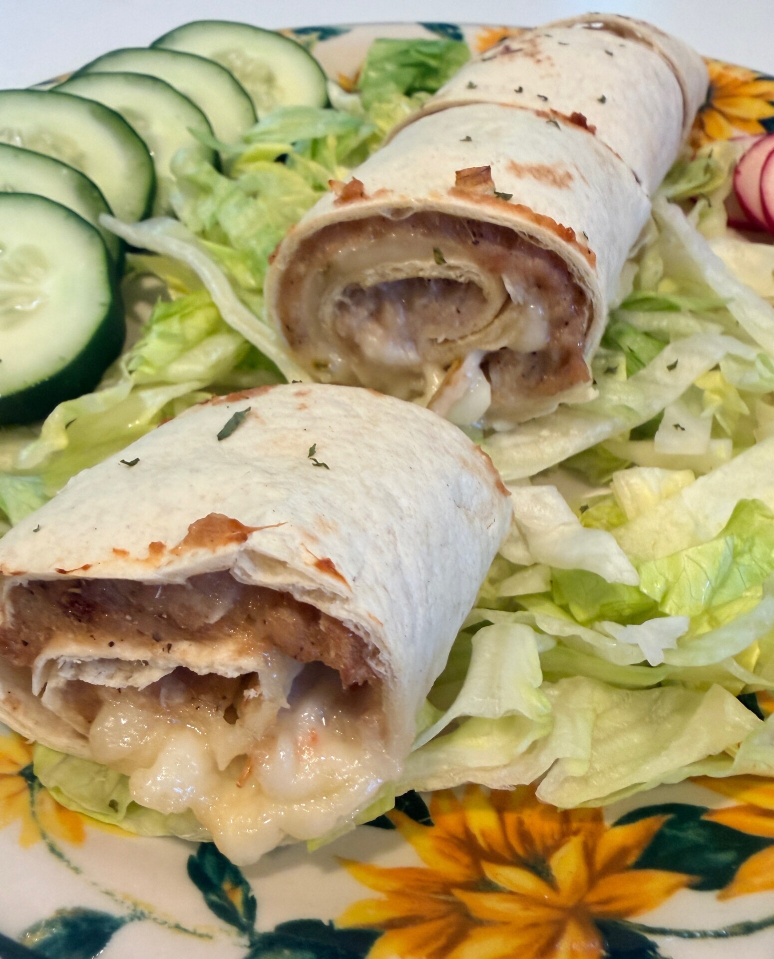 Two cheesy chicken taquitos on a bed of shredded lettuce, served with sliced cucumber and radish on a floral-patterned plate.