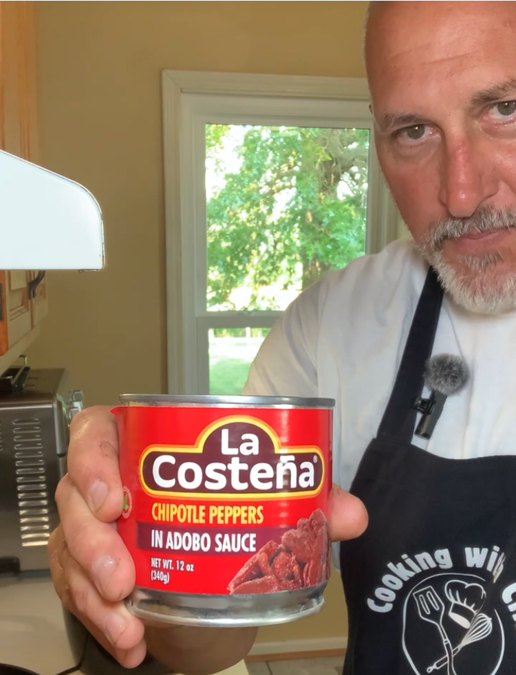A person in a kitchen holds a can of La Costeña chipotle peppers in adobo sauce, wearing a dark apron and white shirt, with a window and greenery in the background.