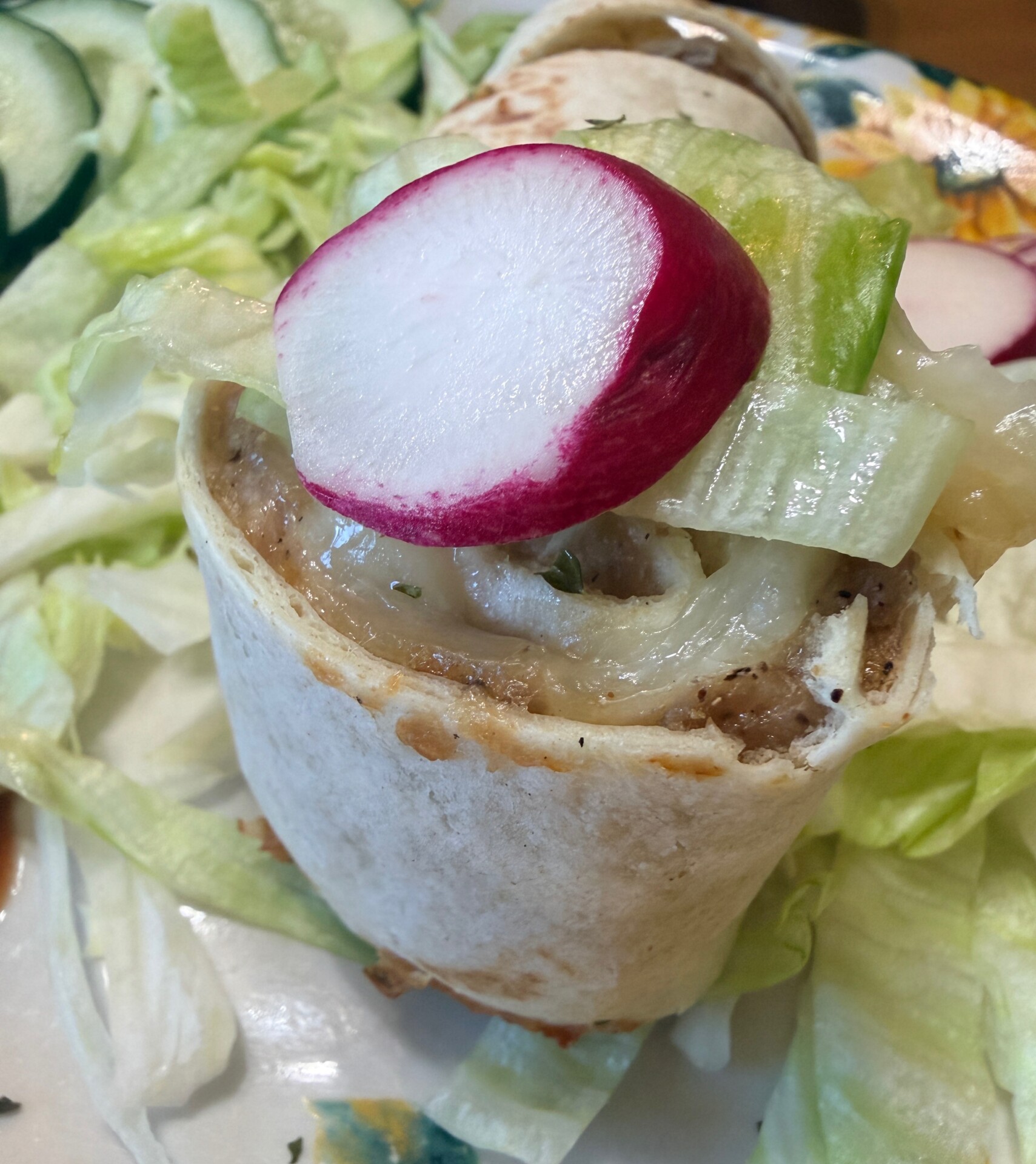 A close-up of a small tortilla wrap filled with seasoned meat, topped with grilled onions, a slice of radish, and lettuce, served on a bed of shredded lettuce.
