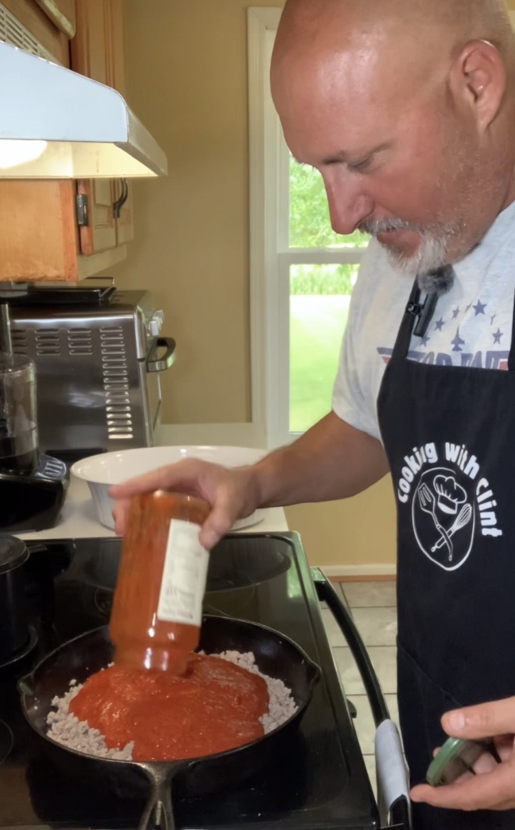 A man wearing a “Cooking with Clint” apron pours tomato sauce from a jar into a skillet of ground meat on a stovetop in a kitchen.