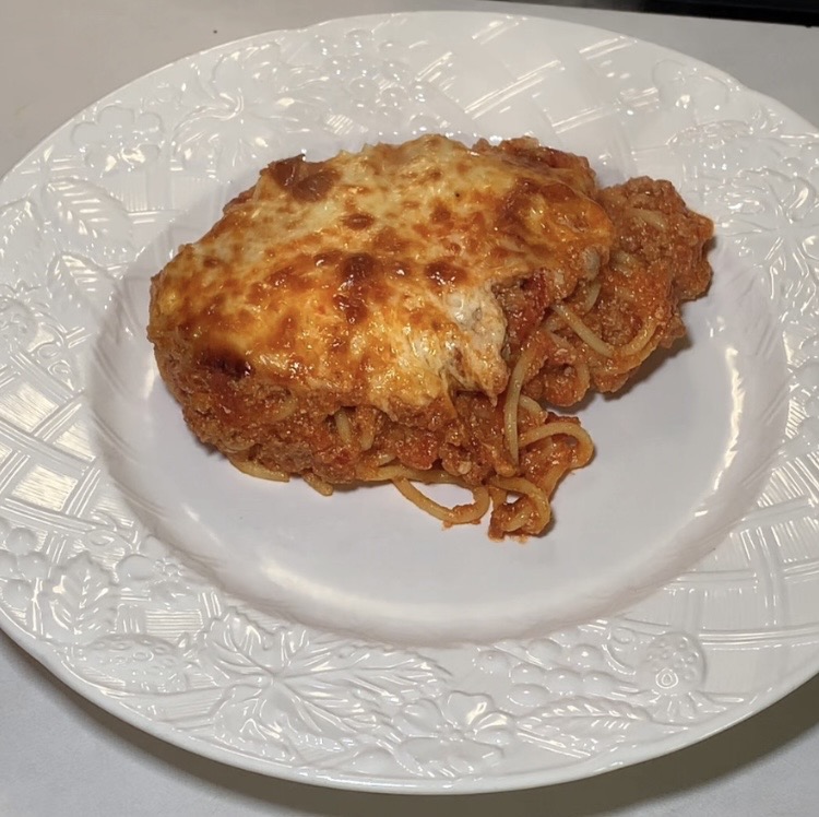 A slice of baked spaghetti with melted cheese and tomato sauce is served on a white decorative plate.