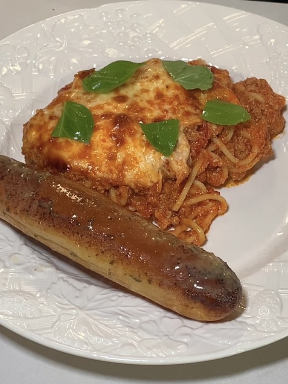 A white plate with cheesy baked spaghetti topped with basil and a browned breadstick.
