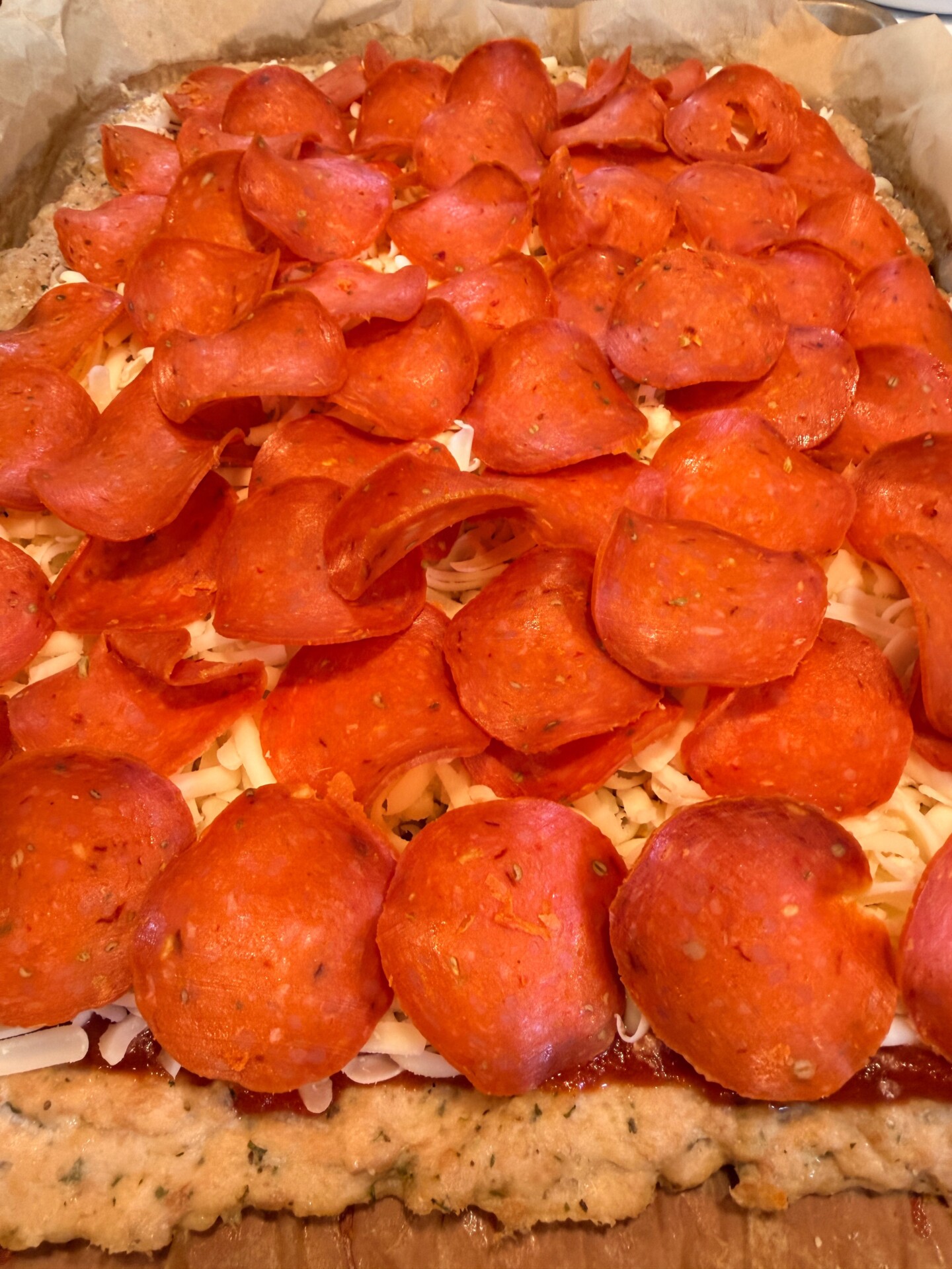 Close-up of a rectangular pizza topped with lots of pepperoni slices and melted cheese on a thick, herb-seasoned crust, shown on parchment paper.
