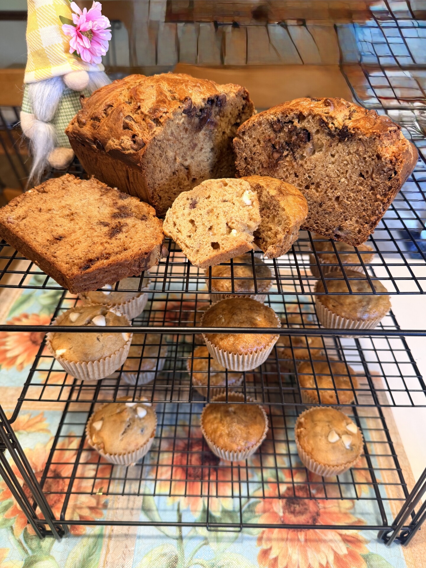 Banana bread loaves and muffins, some sliced to show a moist, nutty interior, displayed on a black cooling rack on a floral tablecloth. A small doll with a flower in its hat is partially visible in the upper left corner.
