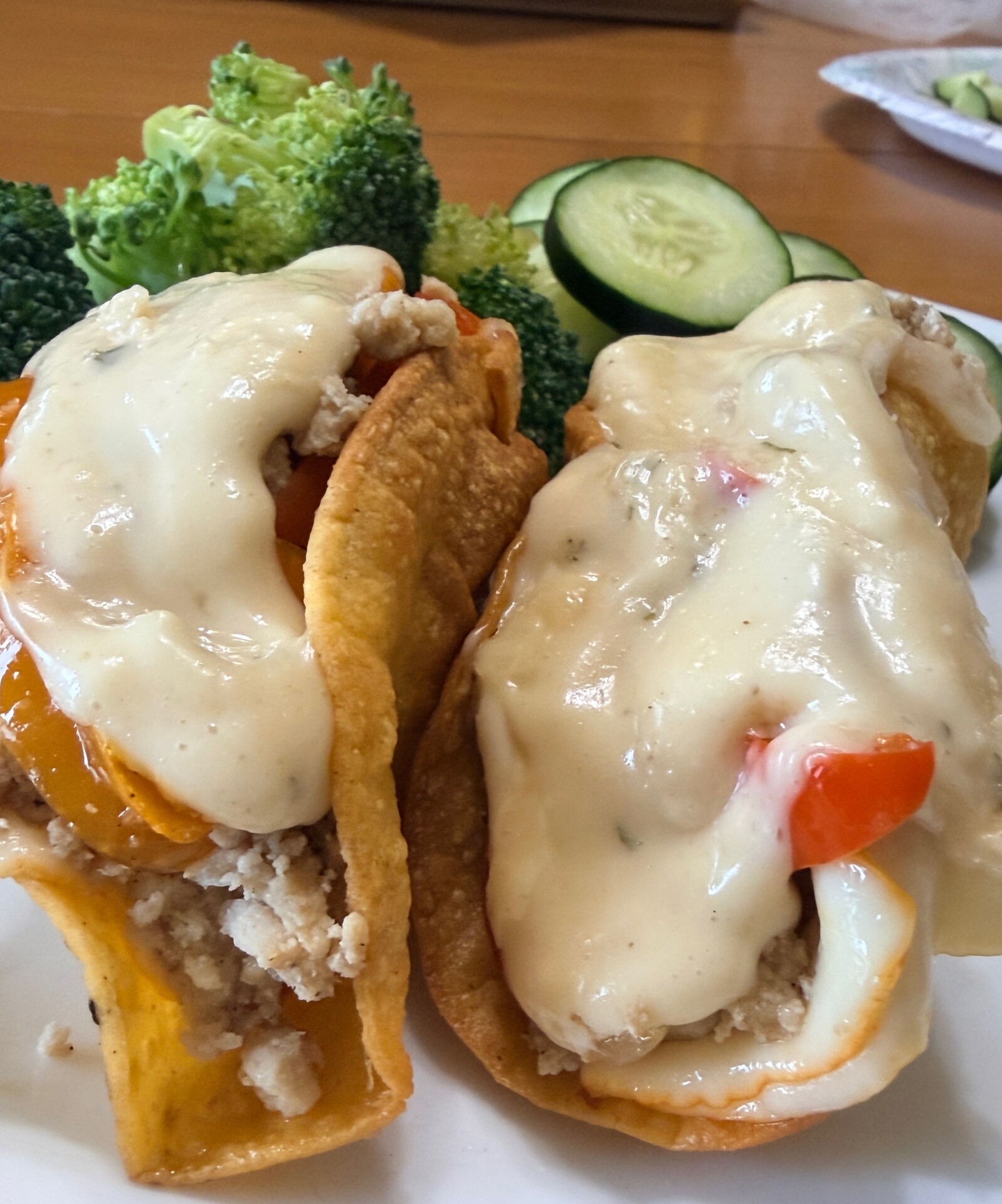 Two stuffed taco shells filled with ground meat, diced peppers, and topped with melted cheese, served with fresh broccoli florets and cucumber slices on a white plate.