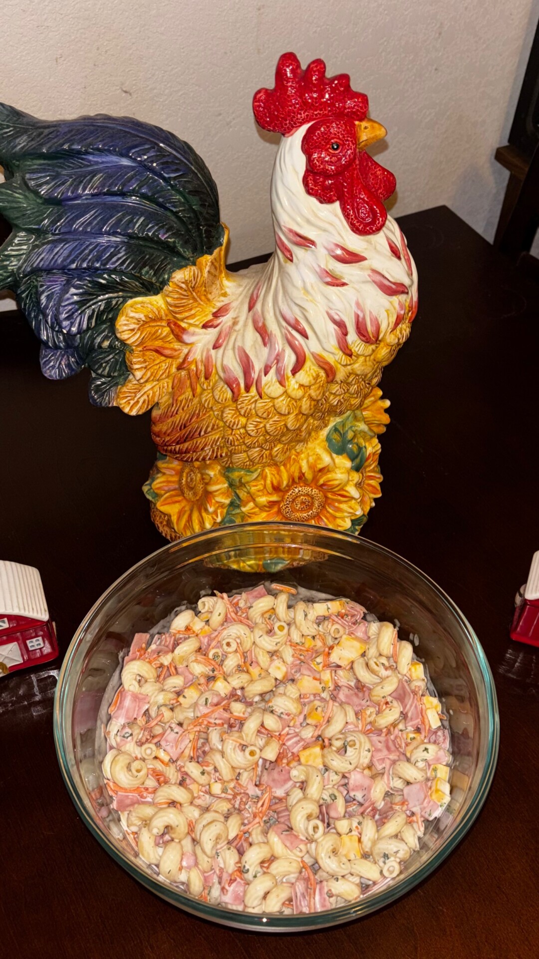 A glass bowl of macaroni salad with ham and cheese sits on a dark table in front of a colorful ceramic rooster statue.