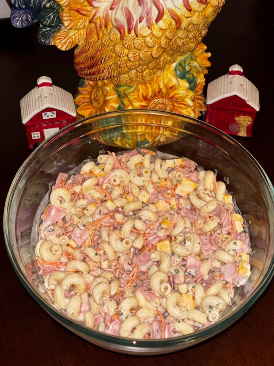 A glass bowl of macaroni salad with ham, cheese, and carrots sits on a table. Behind it are a ceramic rooster and two small red barn decorations.