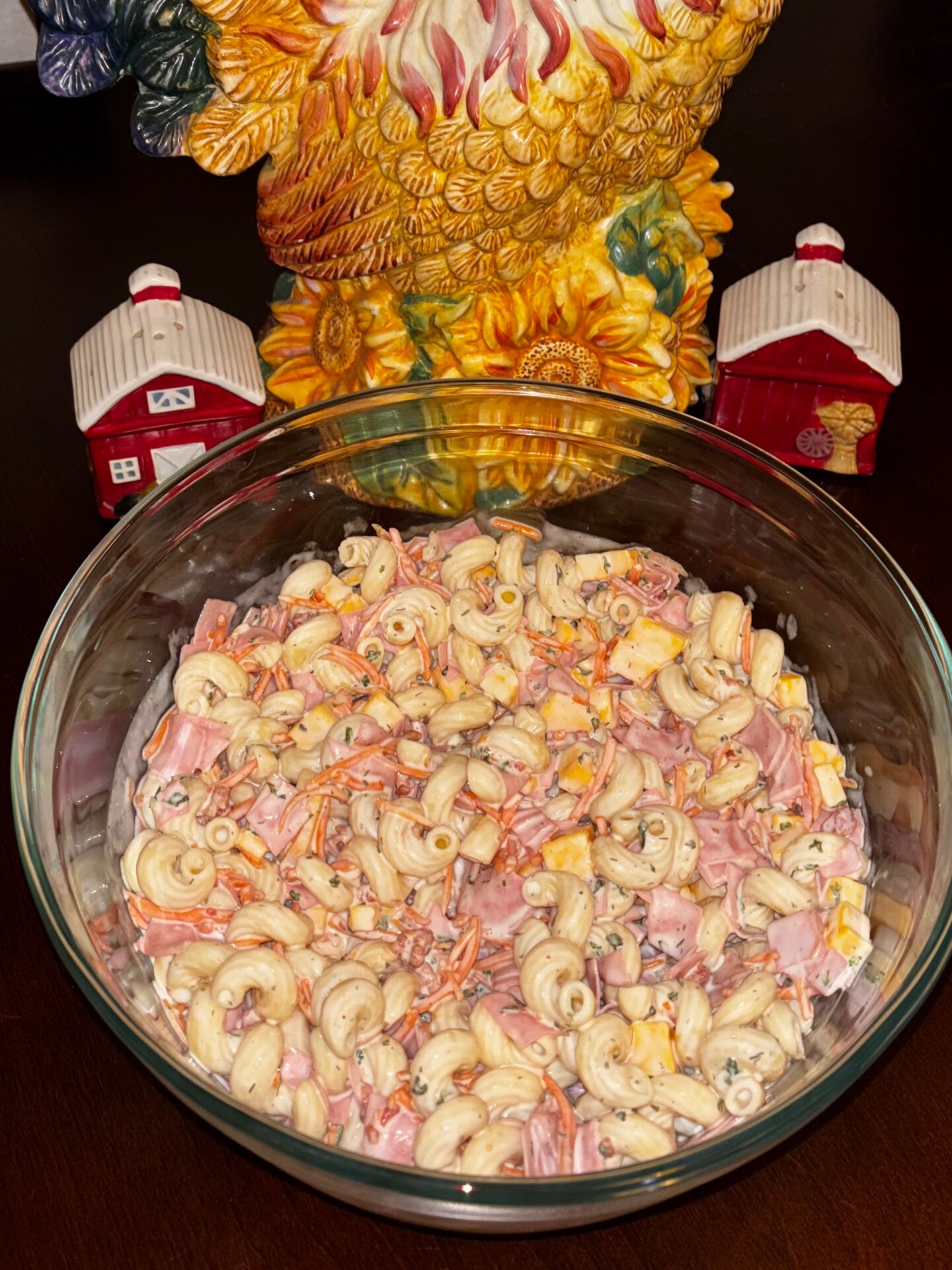 A glass bowl of macaroni salad with ham, cheese, and carrots sits on a table. Behind it are a ceramic rooster and two small red barn decorations.