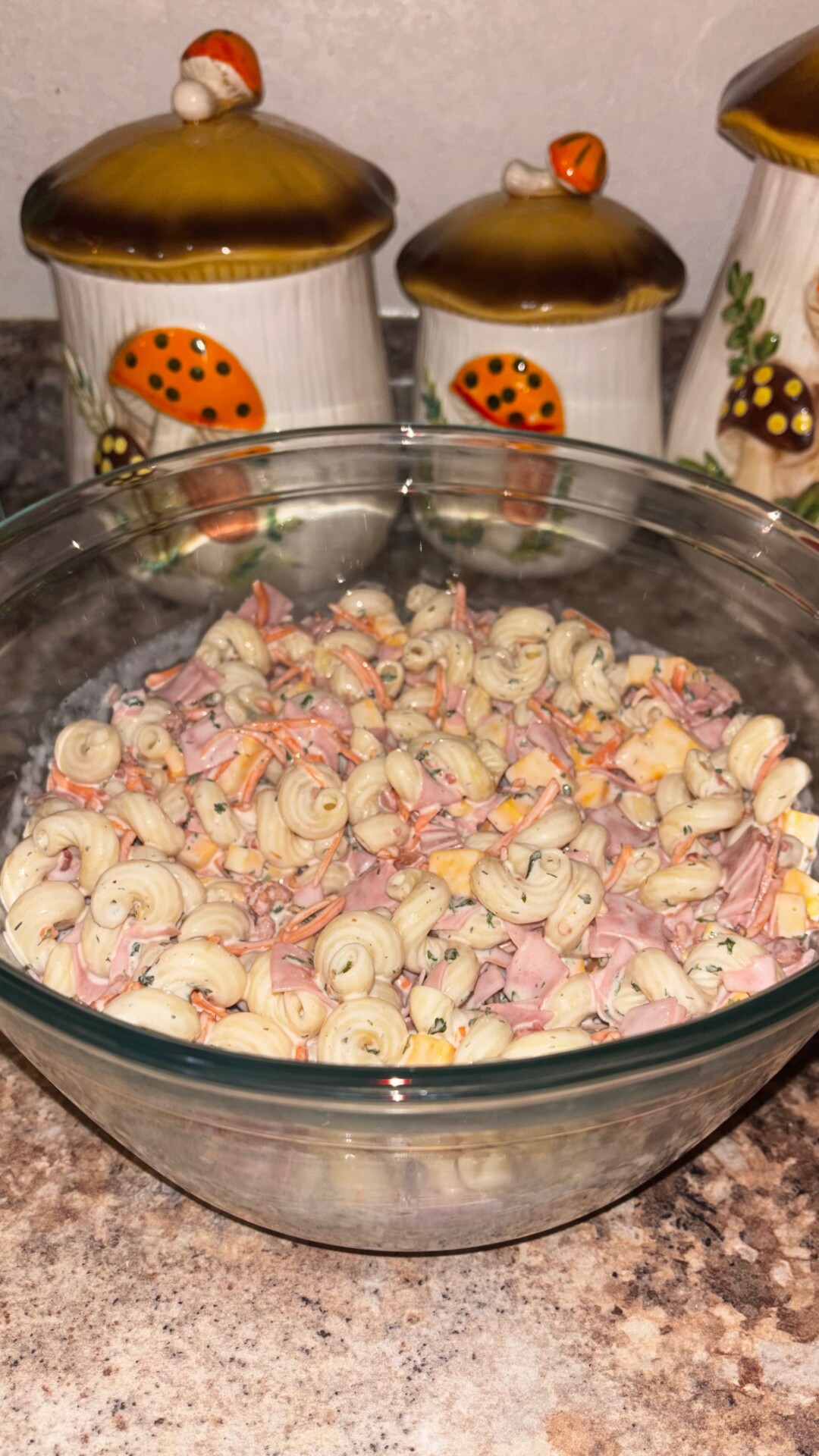 A glass bowl filled with creamy pasta salad containing ham and vegetables sits on a countertop, with three mushroom-themed ceramic canisters in the background.